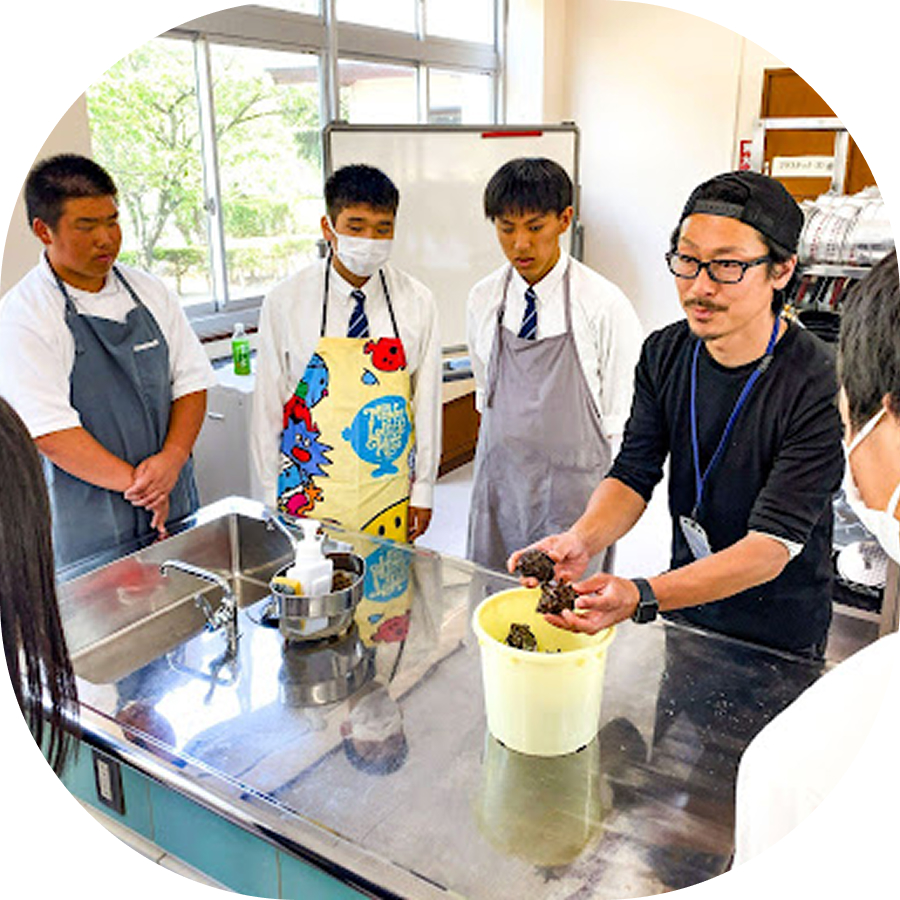 エプロンを着た3人の男子高校生たちが、海産物を持った講師の話を真剣に聞いている様子