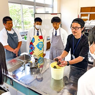 エプロン姿の男子高校生3人が海産物を持った講師に説明を受けている様子
